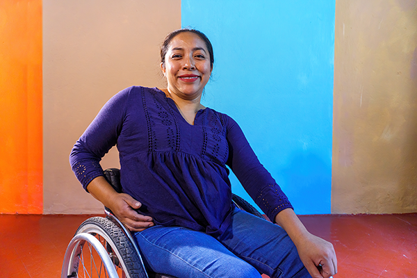 A medium-skinned woman using a wheelchair in front of a brown, orange, and blue background. She has brown hair and is wearing a purple shirt with blue jeans.