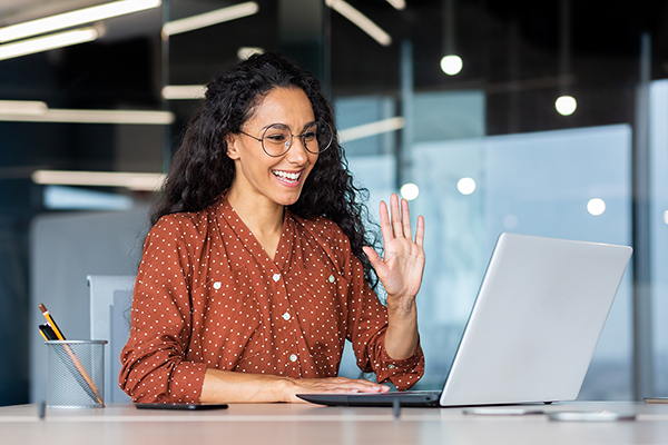 AdobeStock_551563180 alt text: A medium-skinned brunette woman, waving at a silver laptop in a modern office. She is wearing round black, circular glasses, and an orange and white spotted shirt.