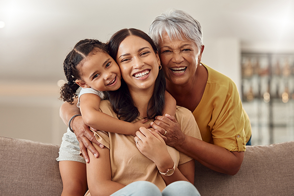 Three medium-skinned women of different age ranges, hugging each other in a living room. On the left is a young girl with dark brown hair, in the middle is a middle-aged woman with dark brown hair, and on the right is an older woman with white hair.